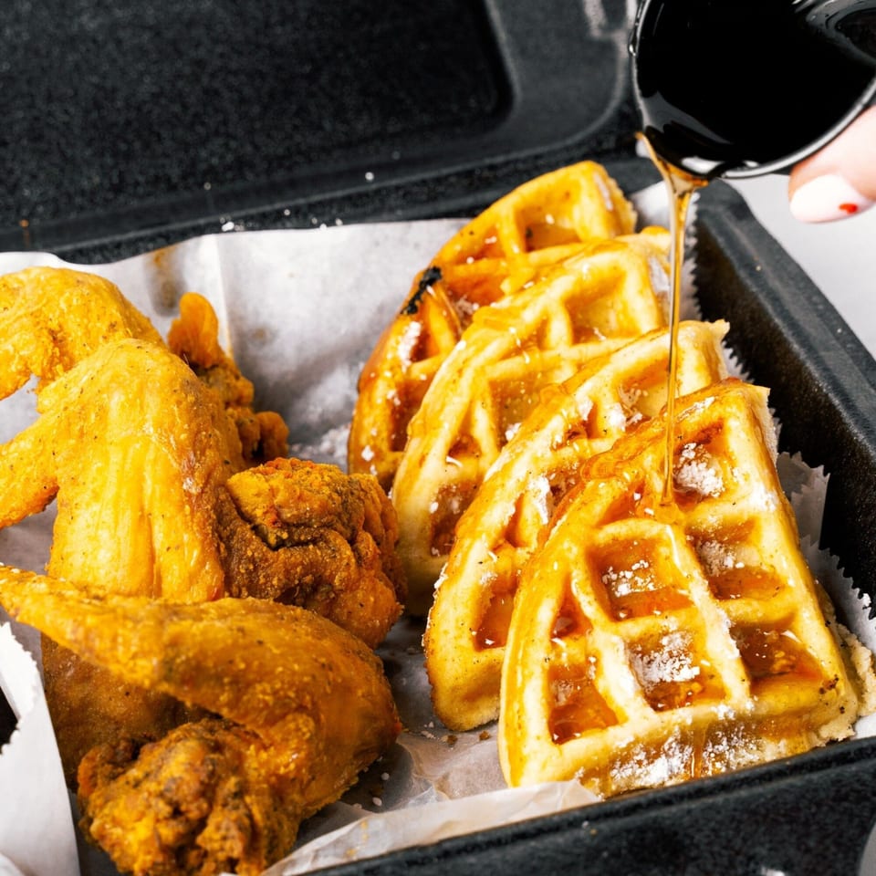 Golden fried chicken wings and waffles with syrup being poured over them in a black takeout container.