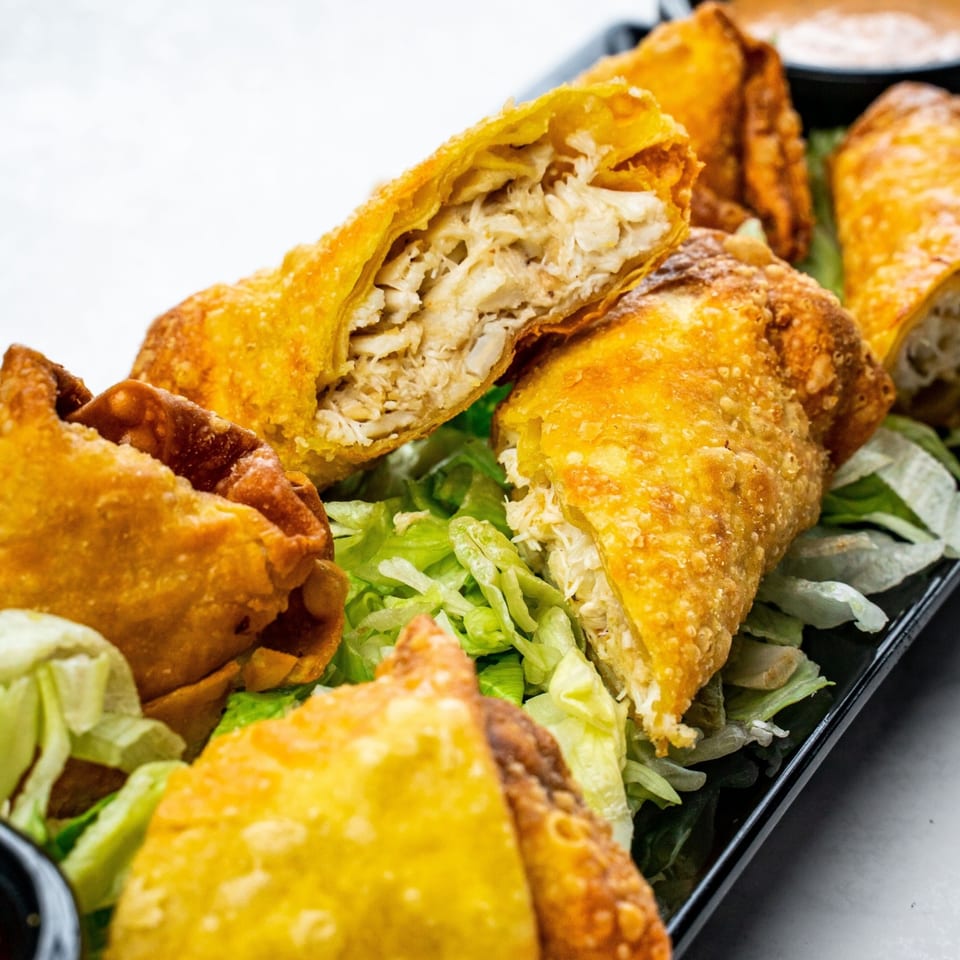 Golden-fried crab rangoon and egg rolls, one cut open to show the crab filling, on a bed of lettuce.